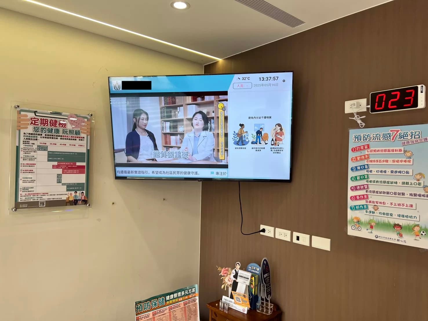 A waiting-room screen mounted on the wall of a clinic, with the ticket display beside it